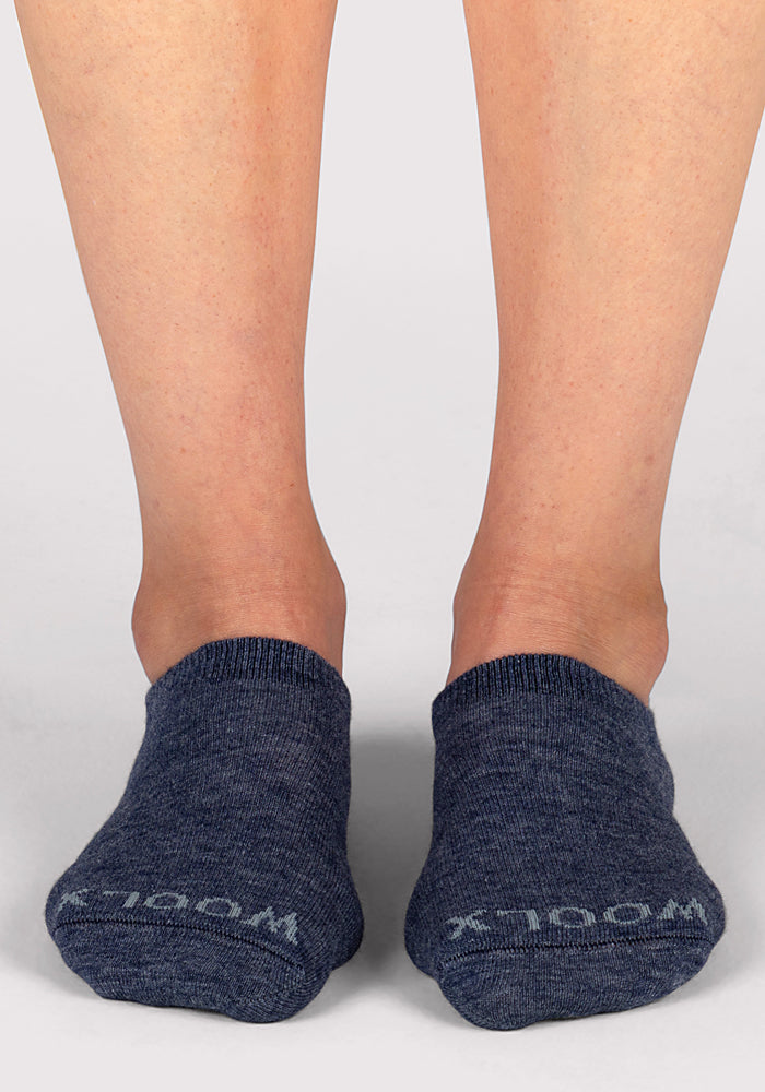 A close-up of someone wearing Woolx Womens Daisy Ped Socks in Blue Jean Dream, standing on a light surface. The socks feature light text near the toes, with only the lower legs and feet visible.