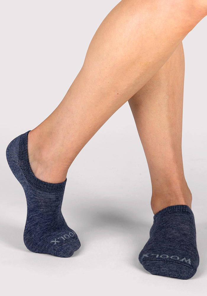 A person stands on a light gray background with legs crossed, wearing Woolx Womens Daisy Ped Socks in Blue Jean Dream, featuring "WOOLX" text on the toes.