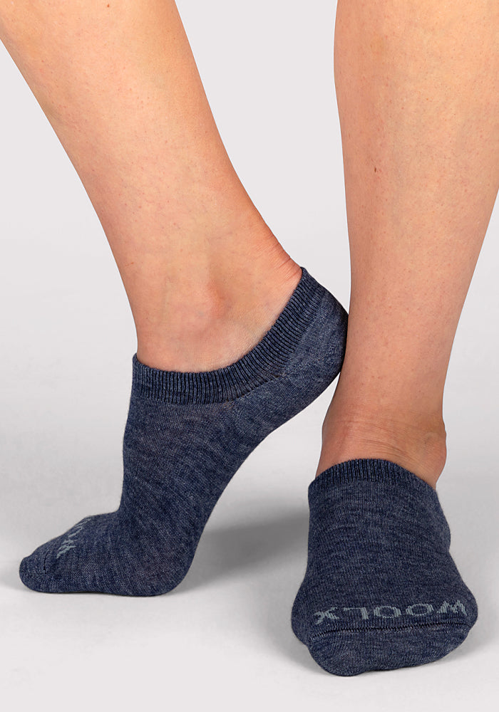 A person wearing blue Women’s Daisy Ped Socks - Blue Jean Dream by Woolx stands on a light gray surface, one foot slightly raised behind the other. The socks feature "Woolx" on the toe.