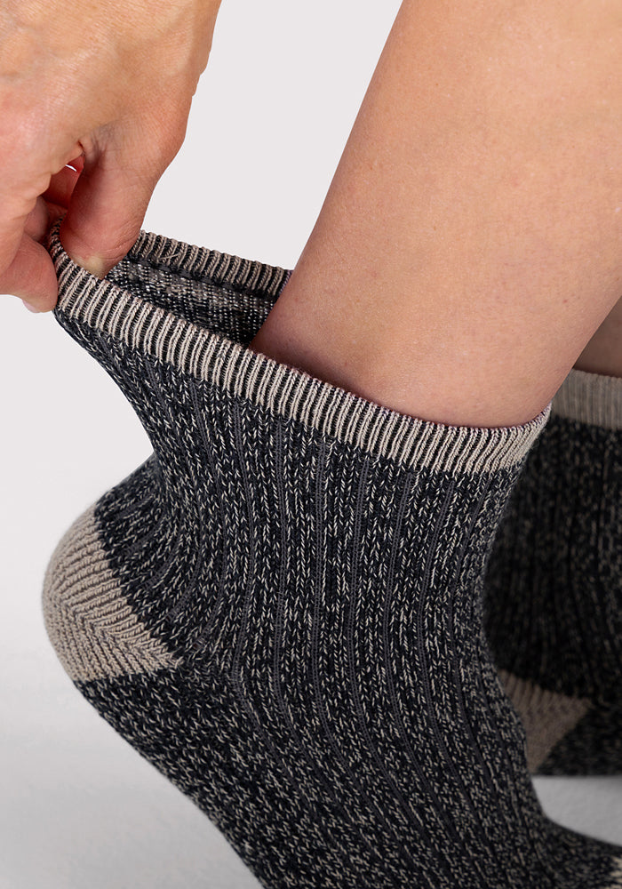 A close-up shows a hand stretching the top of a thick, ribbed Womens Cozy Mini Crew Sock in Black by Woolx, worn on a foot. The luxury sock looks cozy and textured with subtle beige accents.