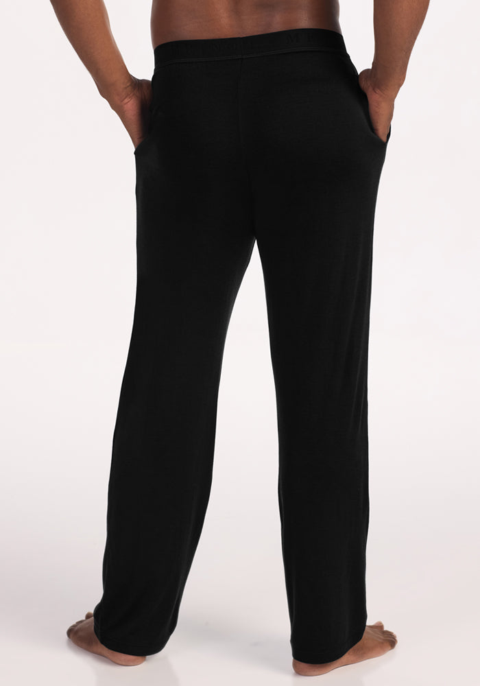 A person stands barefoot on a light surface, hands in the pockets of Woolx Arlo Lounge Pants - Black. The image displays the back view from waist to feet.