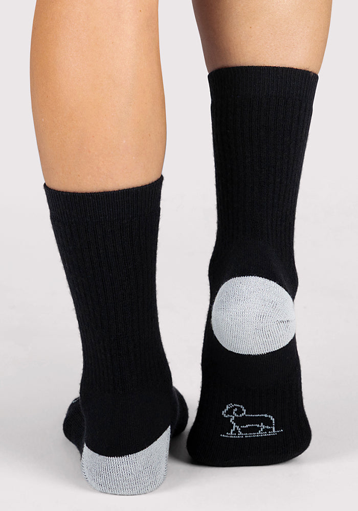 A person wears Woolx Womens Core Crew Socks in black, featuring light gray heels and soles. The cushioned bottom of one sock displays a simple sheep outline. The background is plain and light-colored.