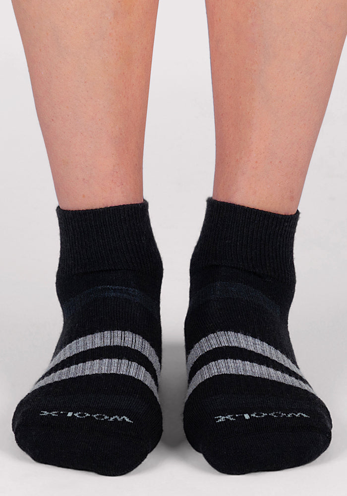 A pair of feet in Woolx Womens Rambler Ankle Sock - Black, featuring two gray stripes and "WOOL" printed on top, stands on a light surface. These Merino wool socks are known for excellent moisture management.