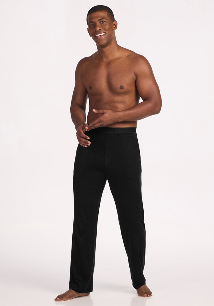 A smiling, shirtless man stands barefoot on a white background wearing Woolx Arlo Lounge Pants in black. He faces forward, with one hand on his waist and the other slightly raised.