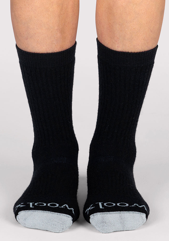 A person wears Woolx Womens Core Crew Socks in black with gray toes and "Wool" in white on each toe. The plain, light background highlights these cushioned hiking socks, perfect for trail comfort.