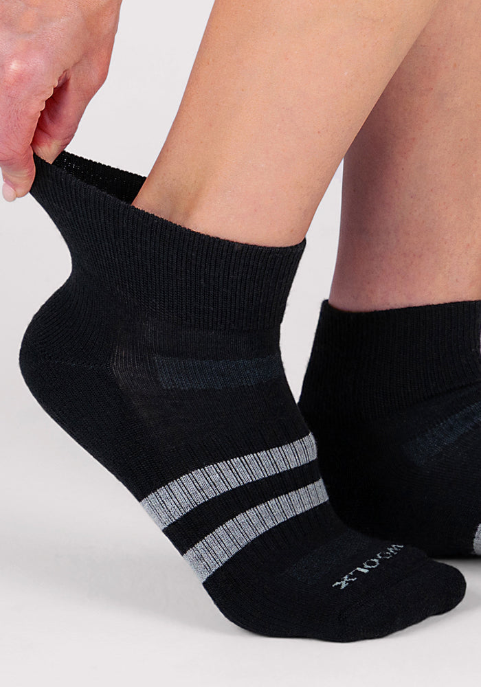 A person is putting on Woolx Womens Rambler Ankle Sock in black, featuring two light gray stripes near the toes, designed for excellent moisture management, against a plain light background.