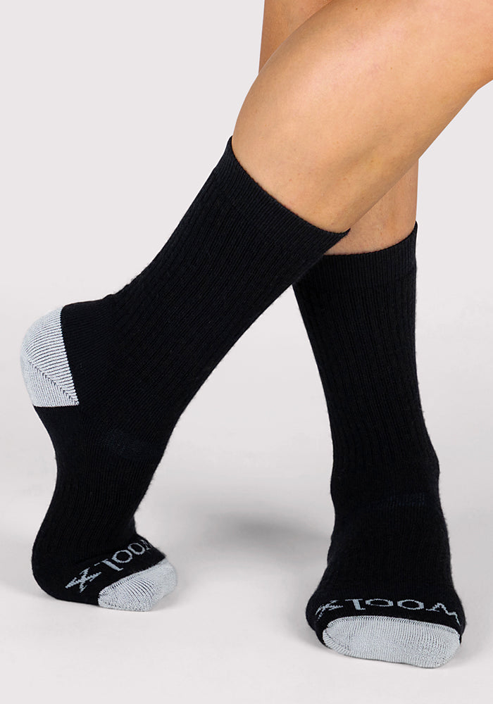 A person wears Woolx Womens Core Crew Socks in black with light gray heels and toes. The socks feature text on the toes and are displayed against a neutral background.