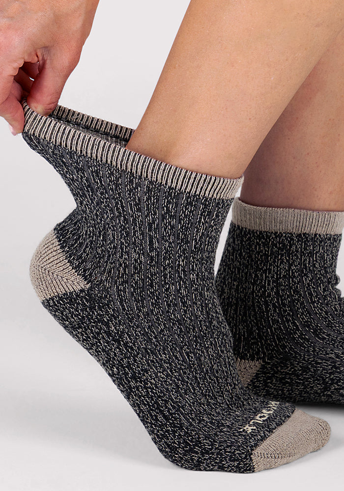 Someone pulls up a Woolx Womens Cozy Mini Crew Sock in black, featuring gray speckles and beige trim. Their other foot already wears the matching sock. The background is plain and light-colored.