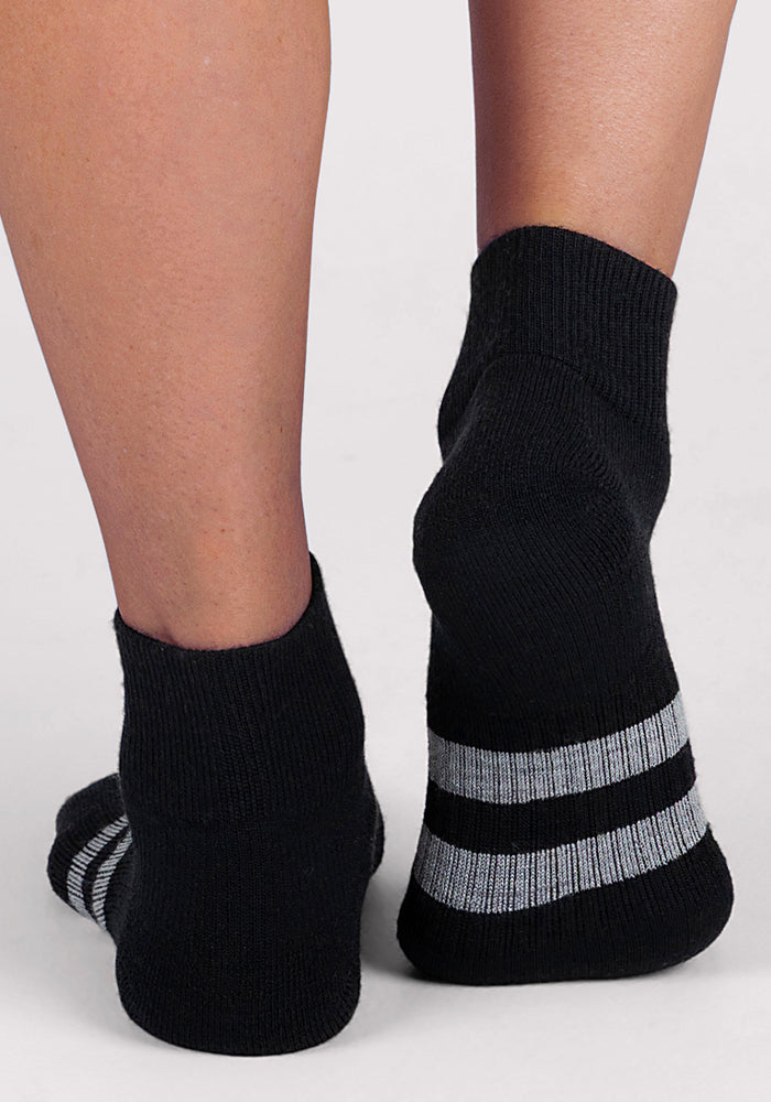 A person stands on a light surface wearing Woolx Womens Rambler Ankle Sock in black with two gray heel stripes, highlighting the comfort and moisture-wicking benefits of Merino wool with legs crossed at the ankles.