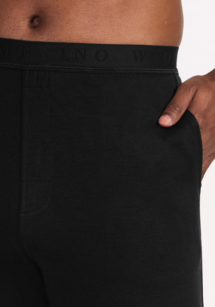 A close-up of a person wearing Woolx Arlo Lounge Pants in black, featuring a visible waistband with subtle lettering and one hand in their pocket, with part of their bare torso showing above the waistband.