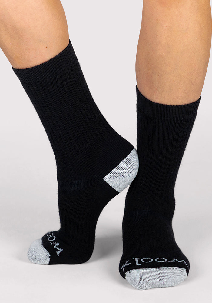 A pair of feet wear Woolx Womens Core Crew Socks in black Merino wool, featuring gray heels and toes, with "WOOL" in white on the toes. The background is plain light gray.
