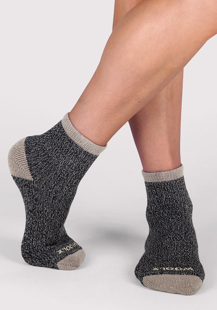 A person stands on tiptoe, showing both the top and side of the Woolx Womens Cozy Mini Crew Sock in black against a plain light background.