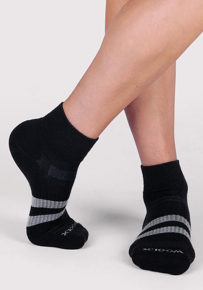 Someone models the Woolx Womens Rambler Ankle Sock - Black, featuring gray stripes, standing with one foot slightly raised against a light background, highlighting the sock’s comfort and moisture-wicking properties.