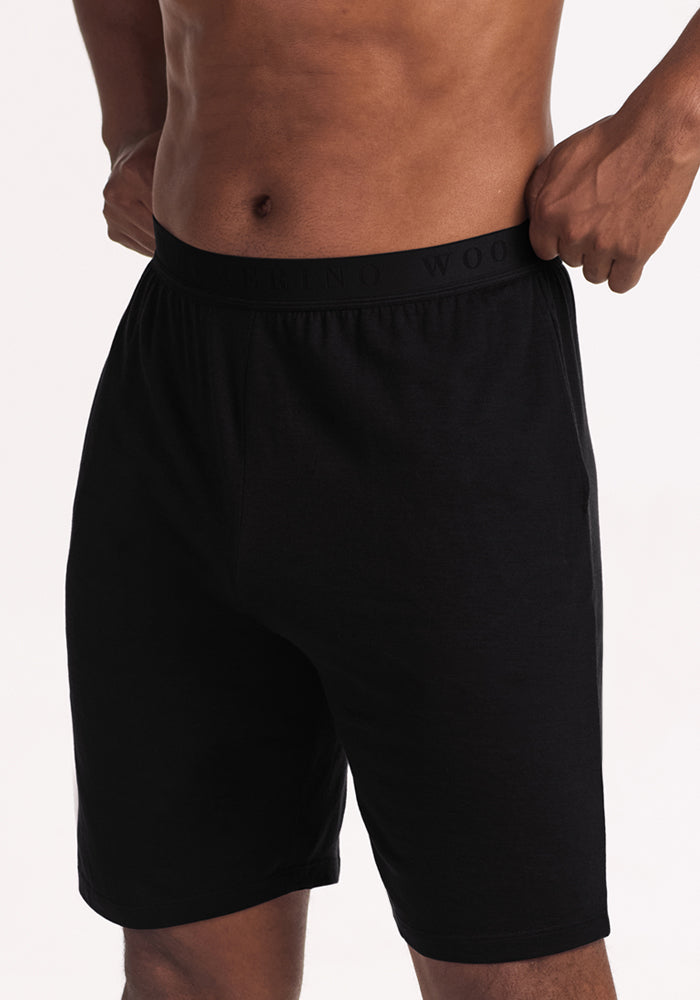 Wearing Woolx Archer Lounge Shorts - Black, a person stands with hands on waistband against a light background.