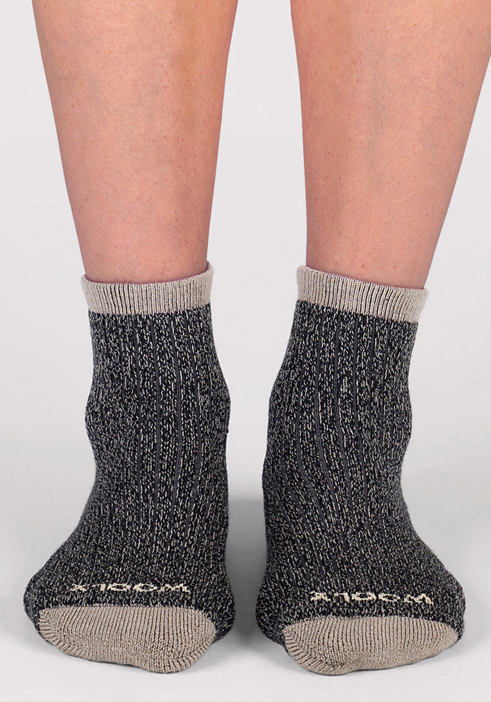 A person wearing Woolx Womens Cozy Mini Crew Socks in black, featuring dark gray speckled Merino wool with light gray toes, heels, and cuffs, stands on a light surface. Only the lower legs and feet are visible.