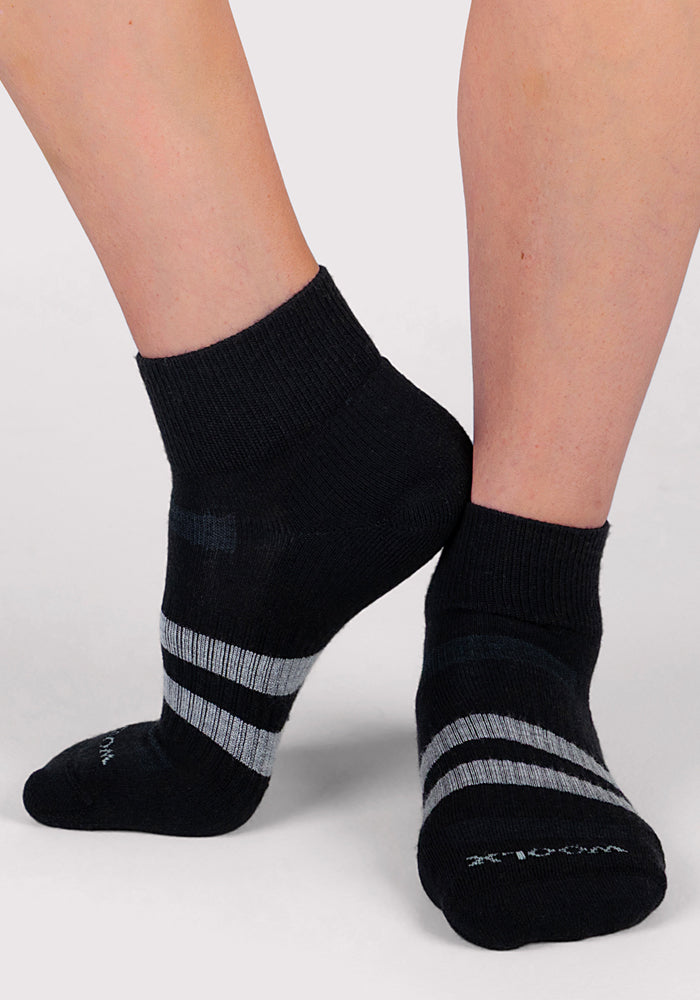 A pair of feet wearing Woolx Womens Rambler Ankle Sock in black with light gray stripes stands on a light background. Enjoy superior moisture management and comfort with every step.
