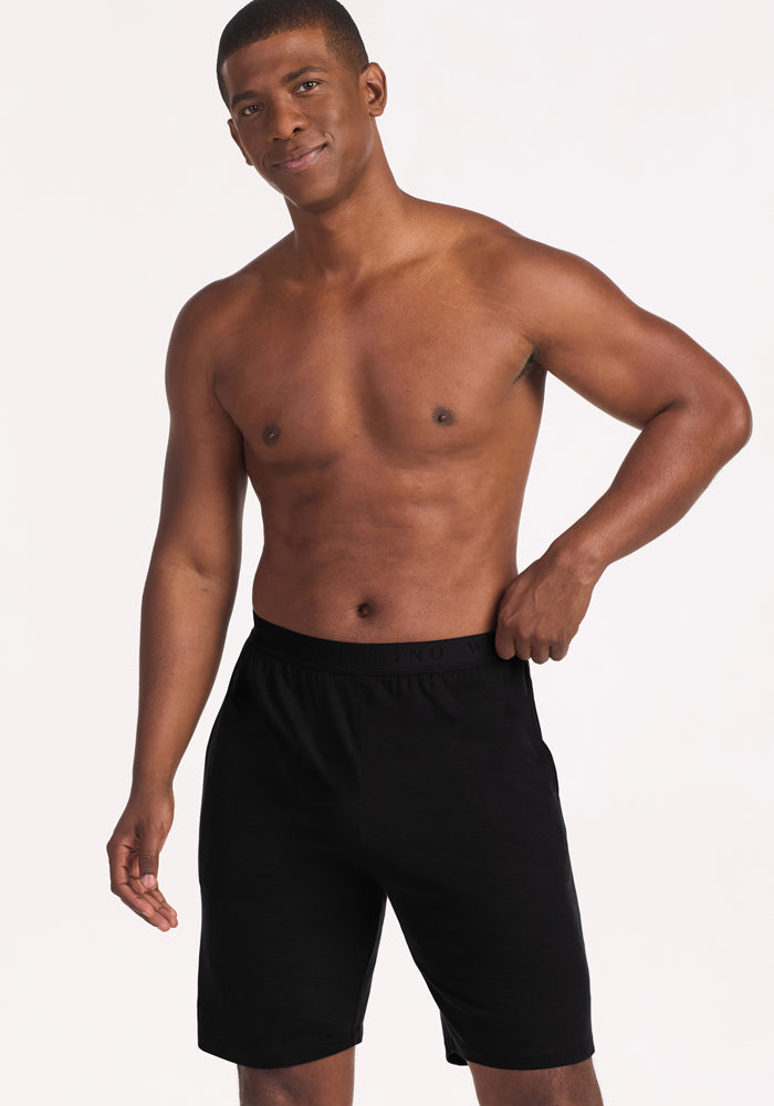 A man smiles, wearing Woolx Archer Lounge Shorts in black, with one hand on his waistband against a plain background.