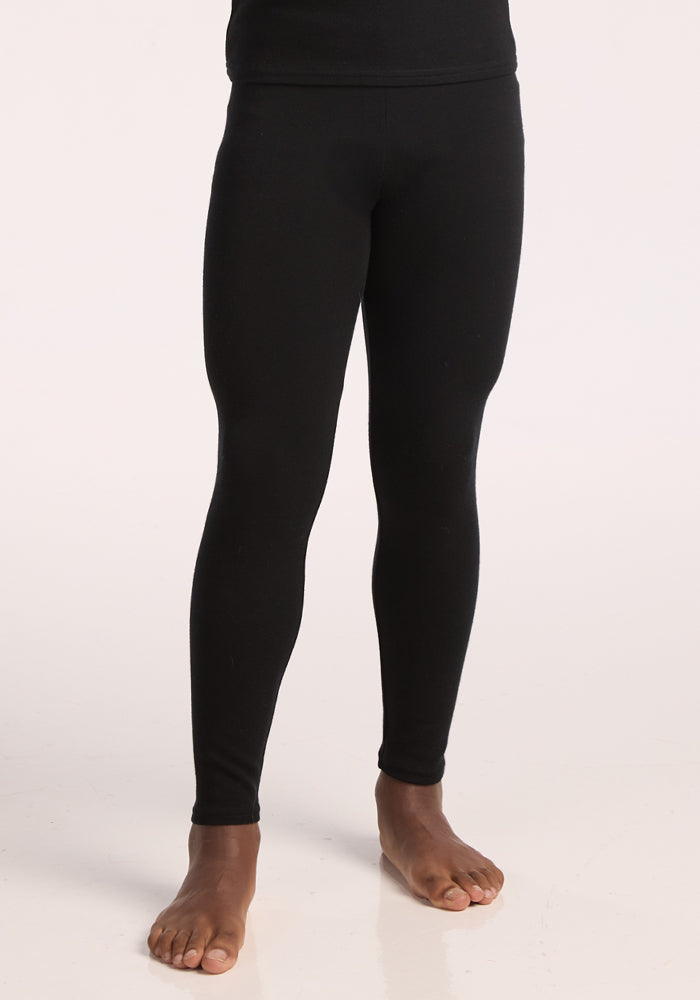 A child stands barefoot in Woolx Kids Explorer Base Layer Bottoms in black, against a light background. The image is cropped from the waist to the ankles. 