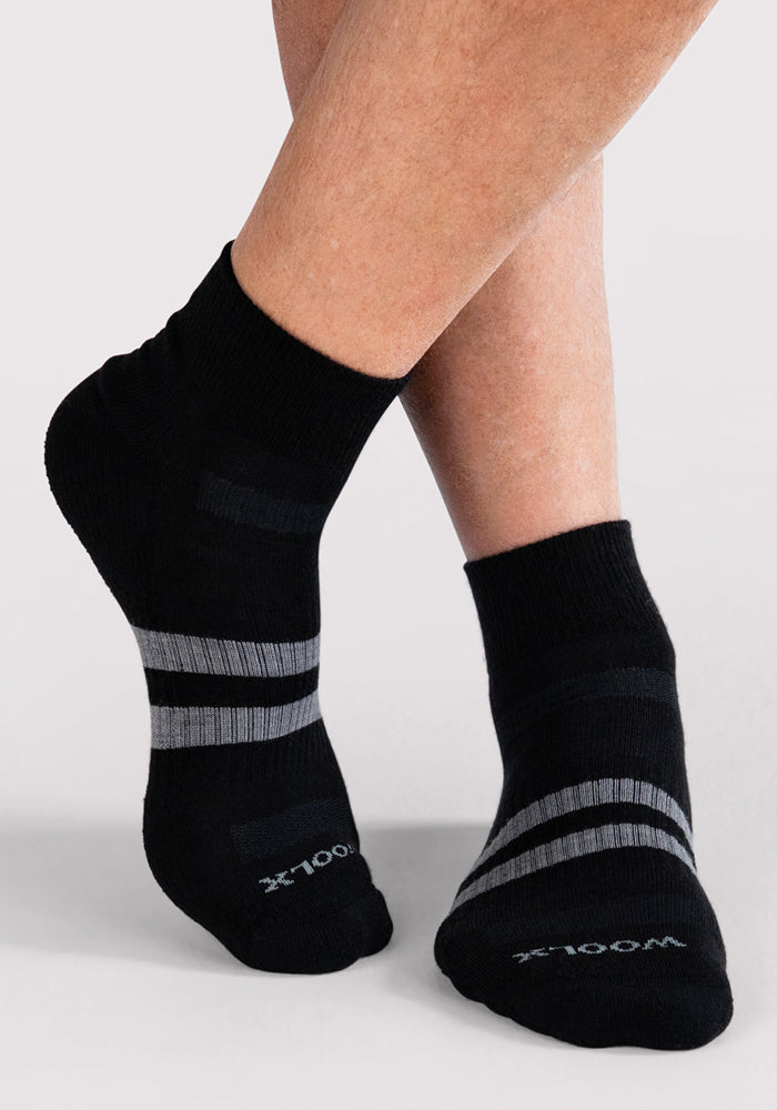 A close-up of crossed legs wearing Woolx Mens Rambler Ankle Sock in black with gray stripes and "WOOLX" on the toes, shown against a plain light background. These socks are known for their moisture-wicking Merino wool construction.