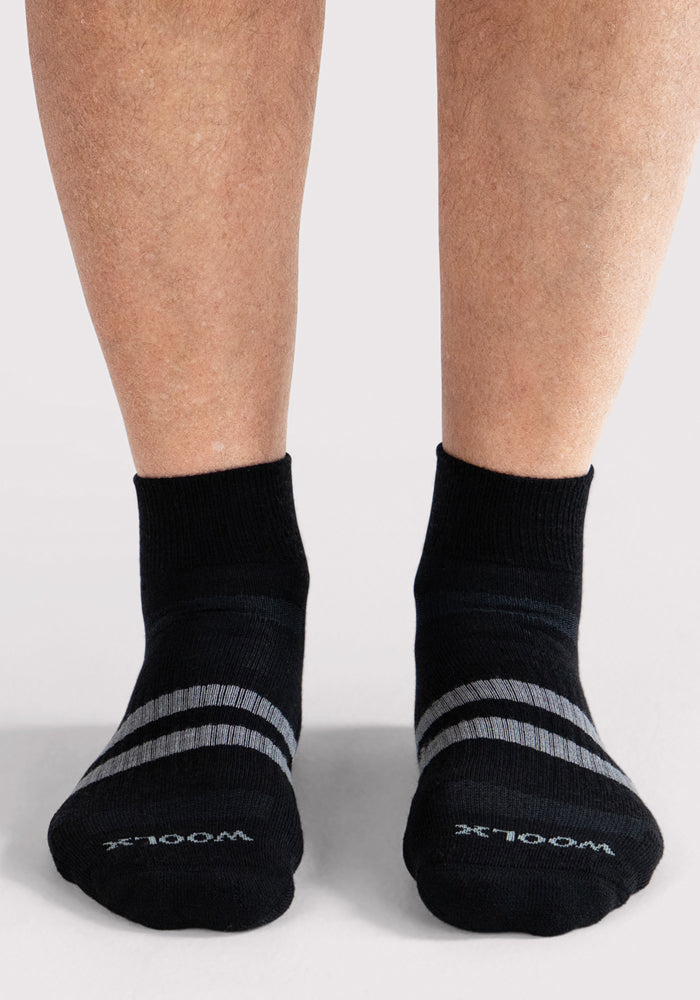 A person wearing Woolx Mens Rambler Ankle Sock - Black with gray stripes and "WOOLX" on the toes stands on a light floor, enjoying comfort and moisture-wicking benefits.