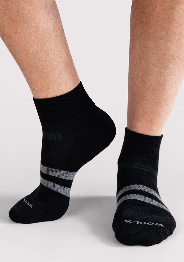 A person wearing Woolx Mens Rambler Ankle Sock - Black, featuring two gray stripes near the toes, stands on a light gray surface. These moisture-wicking socks keep feet dry and comfortable all day.