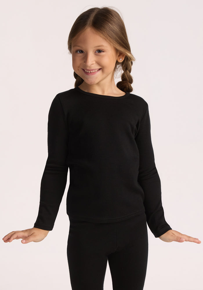 A smiling young girl with light brown braids wears the black Woolx Kids Explorer Base Layer Top. She stands with arms slightly outstretched against a plain light background, showcasing the brand’s moisture-wicking merino wool design. 