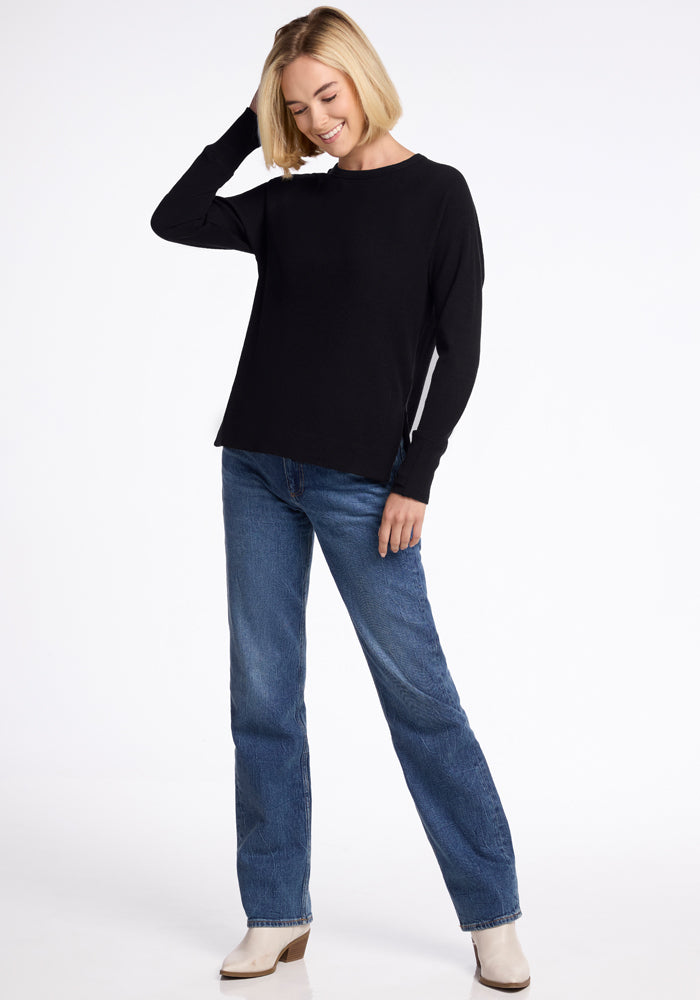 Woman in Woolx Ainsley Petite Sweater - Black, blue jeans, and white boots smiles with one hand on her head.