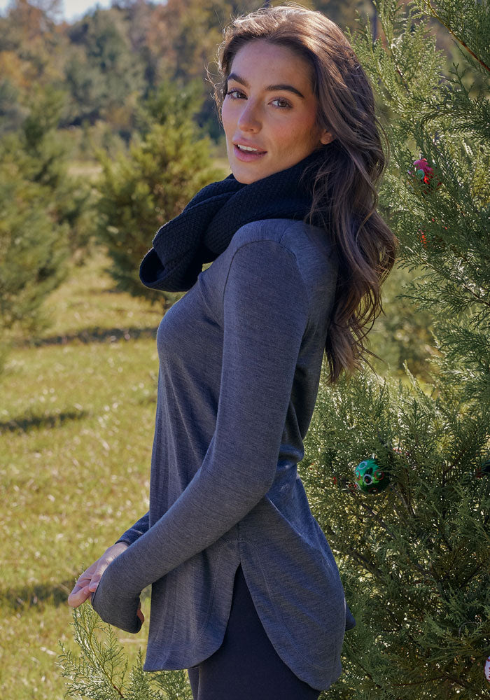 A woman with long brown hair, wearing a gray long-sleeve top and the Woolx Noelle Infinity Scarf in Black, stands outdoors near a small pine tree with greenery and trees in the background on a sunny day.