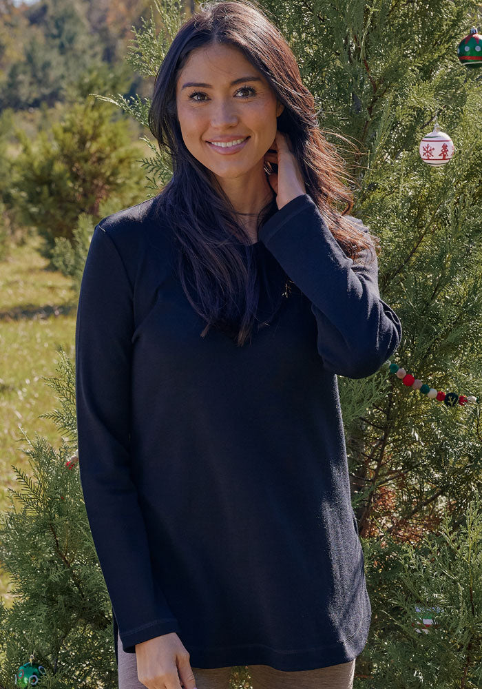 A woman with long dark hair wears the Woolx Blair Tunic in black as she smiles at the camera outdoors beside a decorated evergreen tree on a sunny, grassy day.