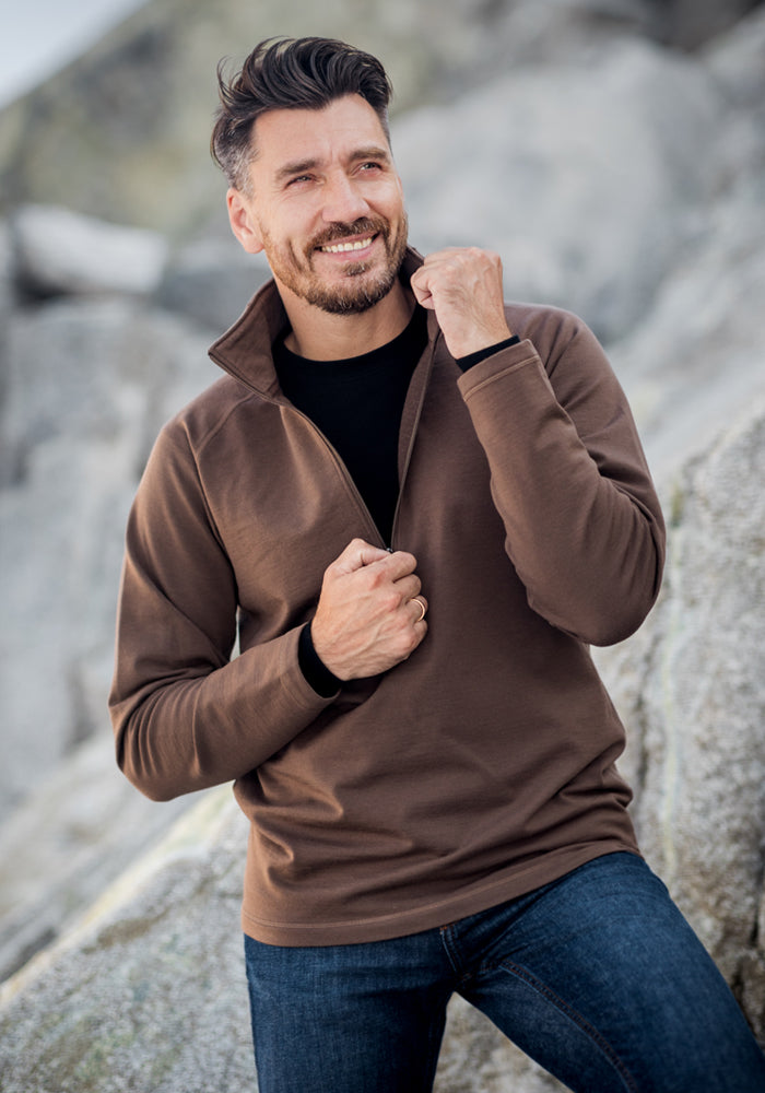A smiling man with short dark hair and a beard is outdoors against a rocky background, wearing the Woolx Blizzard 1/4 Zip brown heavyweight base layer over a black shirt and blue jeans, holding the jacket collar with one hand.
