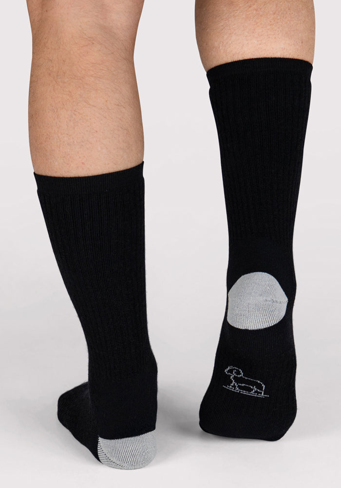 Close-up of a person wearing Woolx Mens Core Crew Socks in black with gray heel and toe accents; the cushioned sole displays a small white dachshund outline.