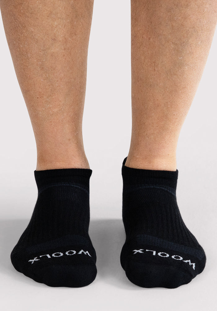 A person wearing Mens Rowan No Show Socks in black by Woolx, featuring "WOOLX" in white on the toes, stands on a light gray surface.