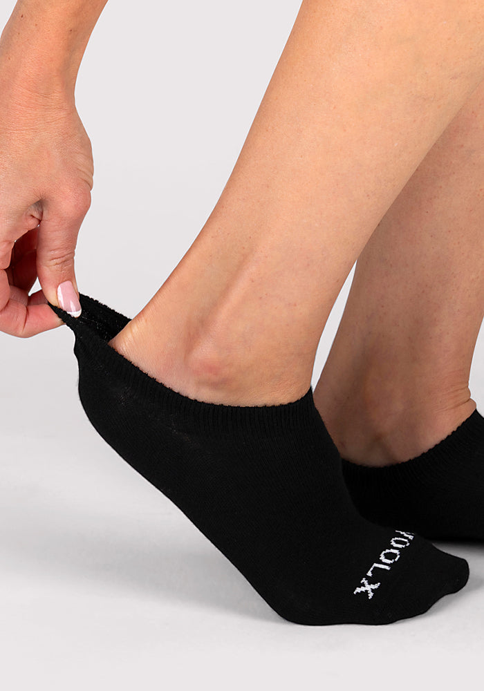 A person with manicured nails pulls up the heel of a Woolx Womens Daisy Ped Sock in black, featuring white text near the toes, showcasing the comfort and style of these women’s socks.
