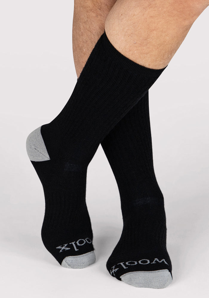 A person wears Woolx Mens Core Crew Socks in black with gray toes and heels. "WOOL" appears in white near the toes. The background is plain and light-colored.