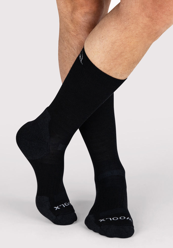 A person stands with legs crossed, wearing Woolx Mens Harbor Crew Socks in black with dark gray accents and white text on the toes, set against a light background.