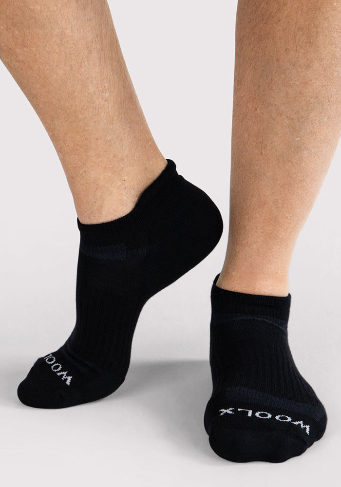A person wears Woolx Mens Rowan No Show Socks in black, featuring "WOOLX" in white on the toes, with one foot slightly raised against a light background.