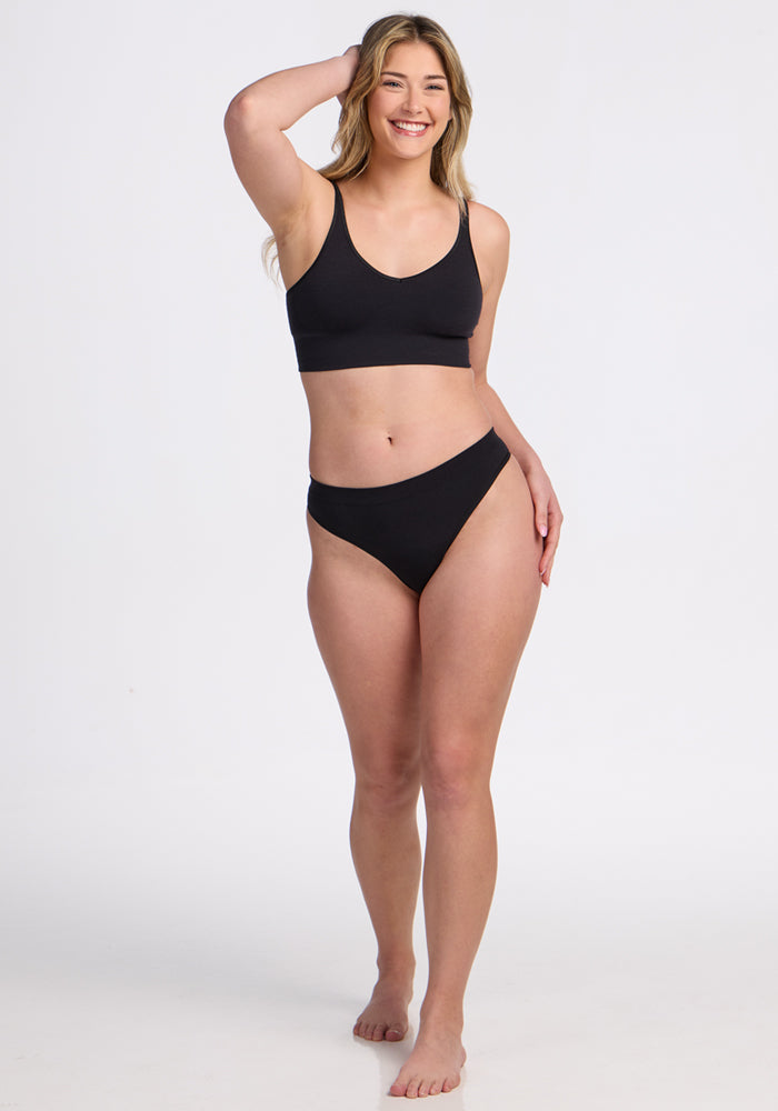 A smiling woman with long, wavy hair stands barefoot against a plain white background, wearing the Woolx Farrah Thong in black and a black sports bra. One hand rests on her head while the other hangs by her side.