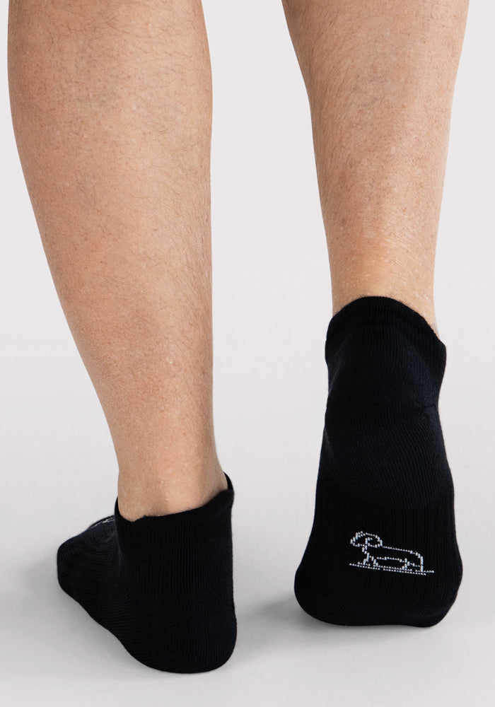 Back view of a person wearing Woolx Mens Rowan No Show Socks in black, featuring a small white dachshund outline on the heel, standing on a light surface.