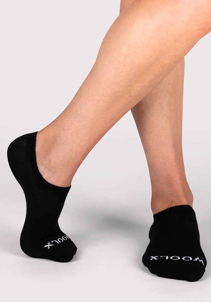 A person with crossed legs models Woolx Womens Daisy Ped Socks in black, standing on a light gray background. The socks add a stylish touch with white “X1000L X” text on the toes.
