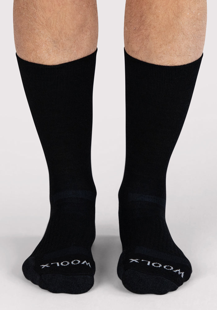 A pair of feet wearing Woolx Mens Harbor Crew Socks in black, featuring "WOOLX" in white lettering on the toes, stands on a light-colored surface.