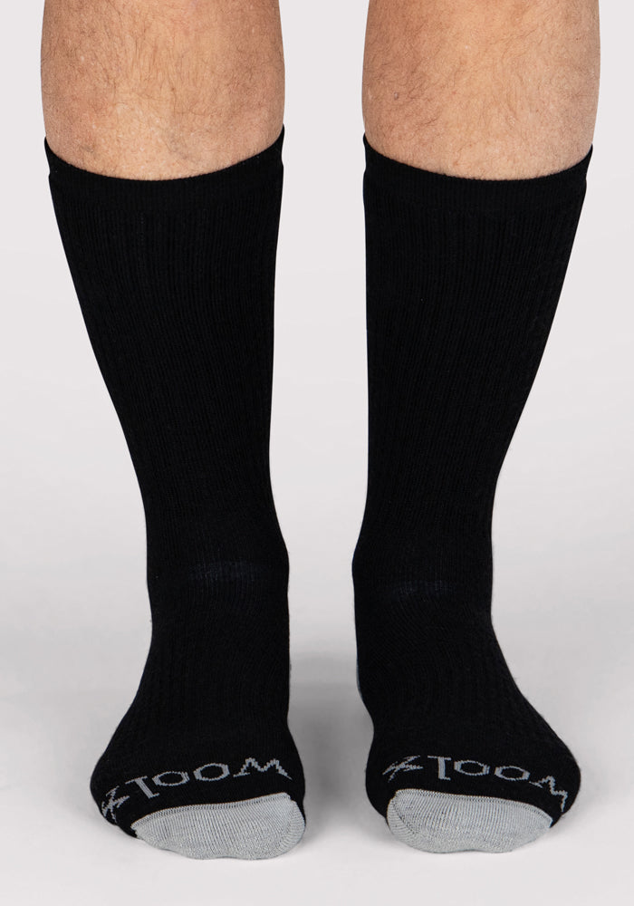 A pair of feet wear Woolx Mens Core Crew Socks in black with gray toes, featuring "ZOOM" in white on the toe. The person stands on a light-colored surface.