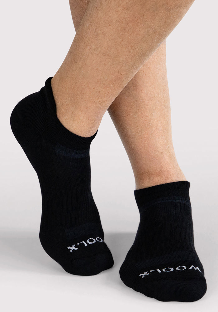 A pair of crossed legs wears Woolx Mens Rowan No Show Socks in black, featuring "WOOLX" in white on the toes, displayed against a plain light background.