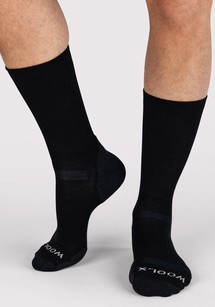 A person wearing Woolx Mens Harbor Crew Sock - Black, featuring "WOOLX" in white on the toes, stands on a light-colored surface. Only their lower legs and feet are visible.