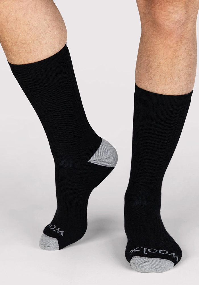A person wears Woolx Mens Core Crew Socks in black with gray toes and heels. "ZOOM" is printed in white on the toes. The background is plain and light-colored.