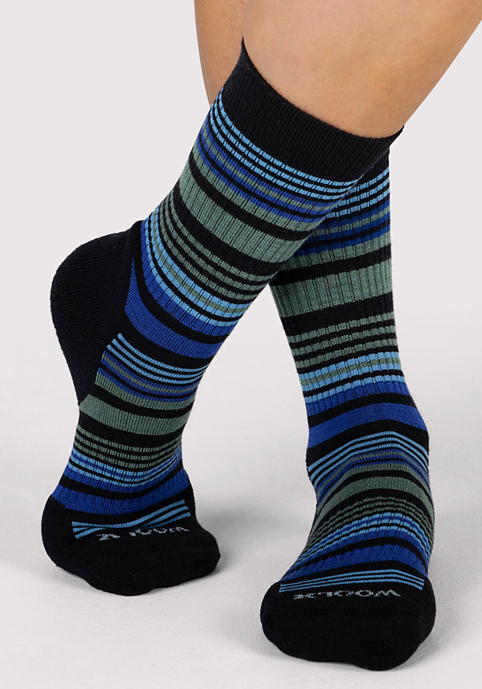 A person stands on a light gray surface wearing Woolx Womens Everyday Crew Socks - Black Stripe, featuring blue, green, and gray horizontal stripes and a ribbed texture for a cozy look, with toes slightly turned inward.