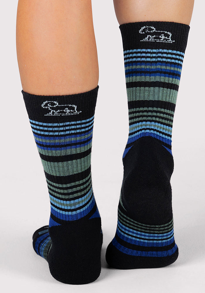 A person wears Woolx Womens Everyday Crew Socks - Black Stripe, featuring black, blue, and green stripes with a small white bunny logo near the top, standing with feet together and heels visible.