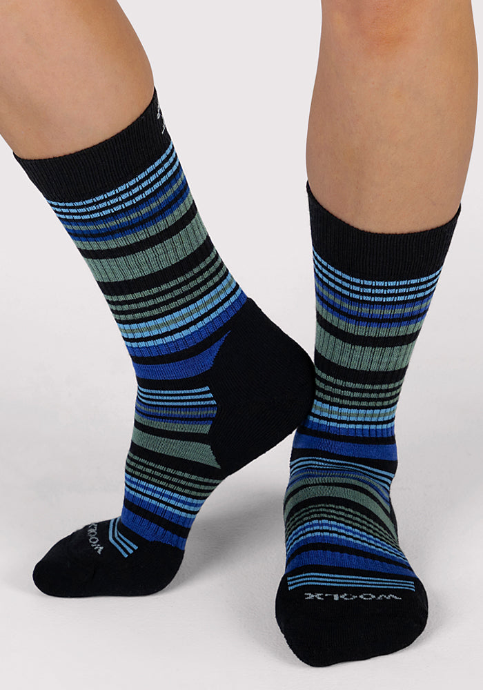 A person stands with one foot crossed behind the other on a light gray background, wearing Woolx Womens Everyday Crew Socks in Black Stripe, featuring moisture-wicking fabric and colorful black, blue, green, and gray stripes.