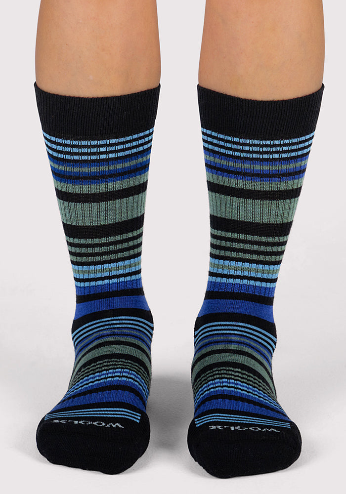 A pair of feet wearing Woolx Womens Everyday Crew Socks - Black Stripe, featuring moisture-wicking fabric and horizontal stripes in blue and gray, stands against a plain white background.