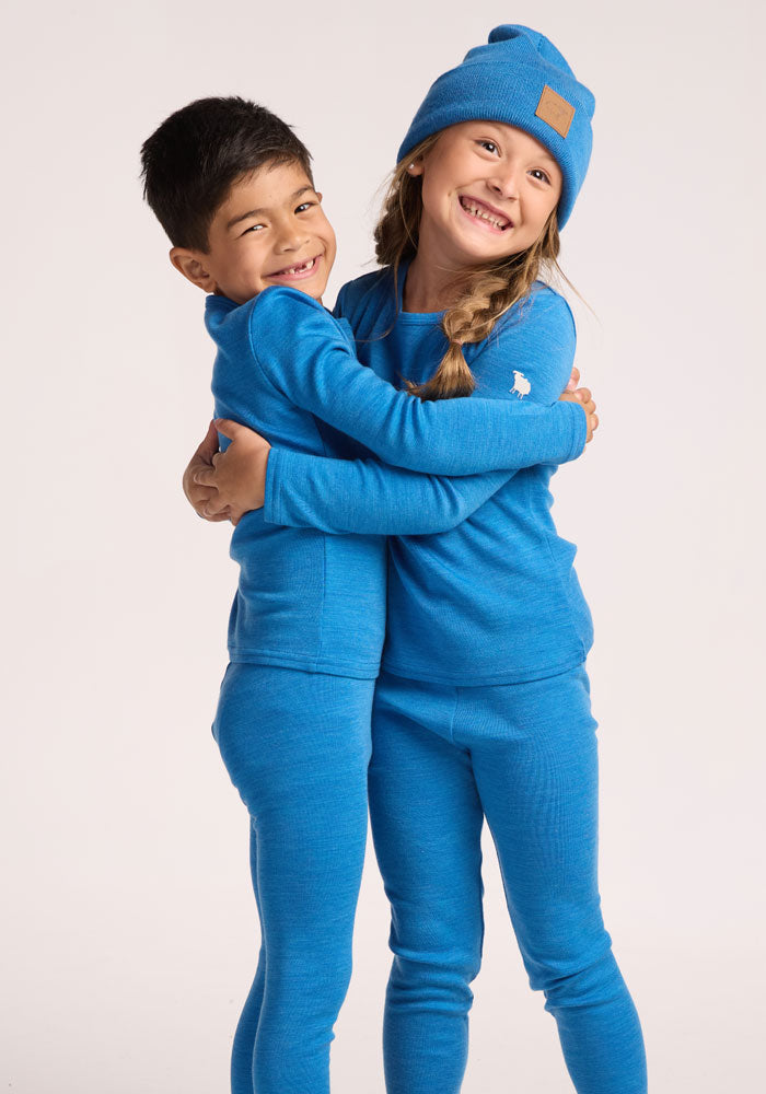 Two smiling children hug, both wearing Woolx Kids Explorer Base Layer Bottoms in Blue Razz. The girl also sports a blue beanie. They look happy, facing the camera against a plain white background.
