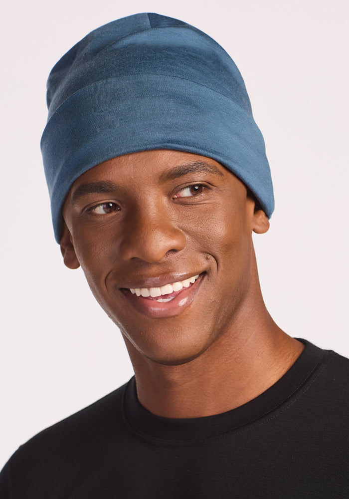 A smiling man models the Woolx Journey Hat in Steel Blue and a black shirt, looking slightly to the side against a plain light background, highlighting the comfort and moisture-wicking benefits of Australian Merino wool.